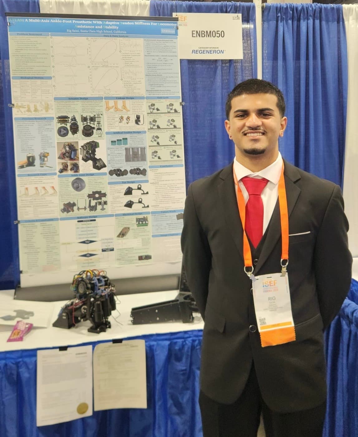 Rig Saini standing beside his ISEF project board for ATLAS, a multi-axis ankle-foot prosthetic, with the prosthetic prototype on the table in front of it.